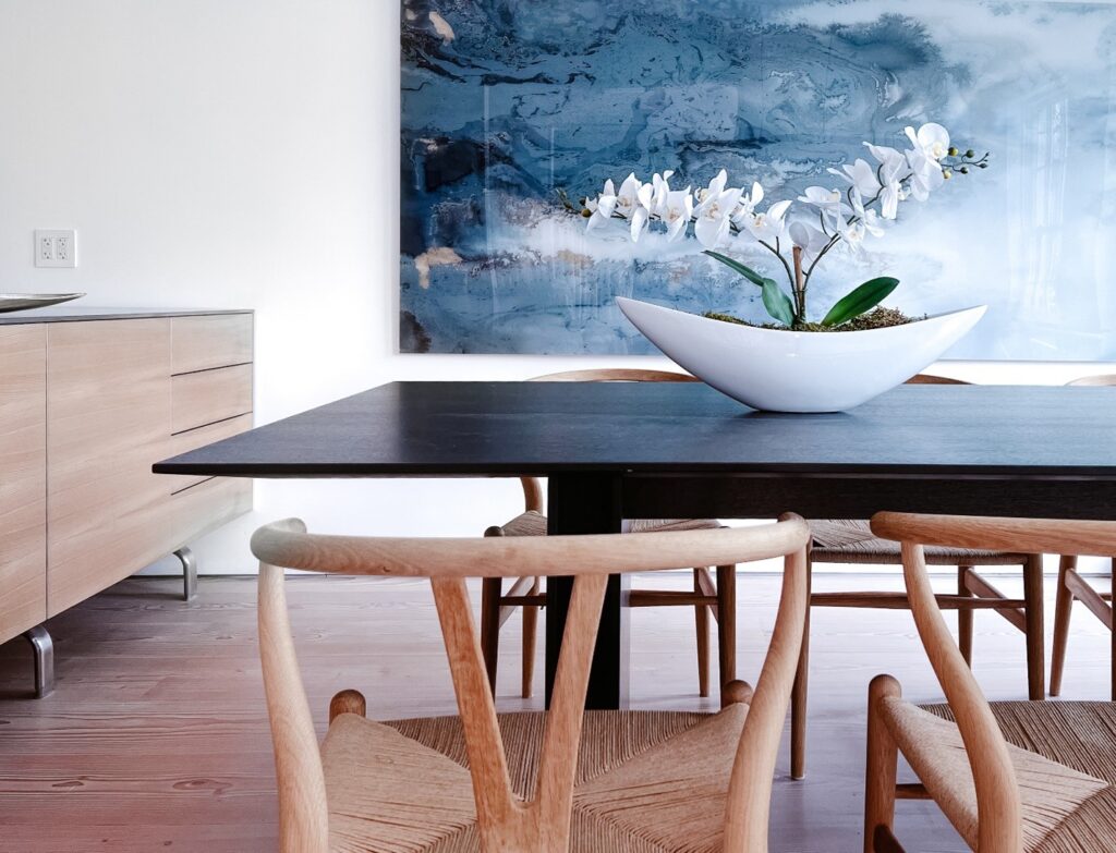 This after photo showcases how an extra-large plexiglass artwork perfectly completes the room, thanks to its commanding presence. The theme of contrast is evident throughout, highlighted by the sleek, modern black wood table and the minimalist wishbone chairs. The artwork not only enhances this theme but also elevates the room's aesthetic beyond what the furniture alone achieves. Artwork of this scale demonstrates that it can be the focal point of a room, doing the bulk of the aesthetic work, which allows the other walls to remain minimally adorned.